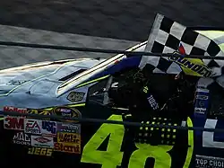 Jimmie Johnson holding a checkered flag in his left hand outside a left-hand side race car window