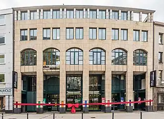Four-storey building of an 80s department store with a light-colored quartzite façade. The first floor with its arches is "closed off" by a large red ribbon