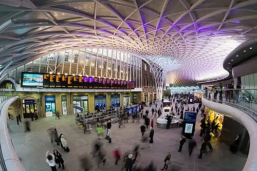 A panoramic view of the inside of King's Cross station. The photo is taken from an elevated position above the concourse, where motion-blurred people walk through and some stand looking at LED screens mounted on the walls