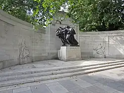 A memorial in white stone, with a central bronze sculpture of a woman, accompanied by a boy and a girl carrying garlands of flowers