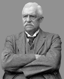 A portrait of a stern-looking man with white hair. He is wearing a suit and has his arms crossed over his chest.