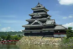 A large five-story castle tower with black wooden walls located on a platform of unhewn stones surrounded on two sides by water. The tower is connected to the lower structure.