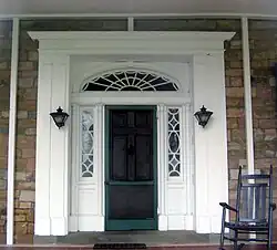 A green door in a white frame with a semi-circular window above with spider-web like rays. On either side of the door are windows with elaborate frameworks, along with two black lanterns, and stone walls. A rocking chair is at right.