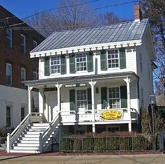 A two-story white wooden house with decorative touches, a pointed metal roof and green shutters. A sign in the center of the porch reads "Irvington History Center" while another at the right on the story below says "Frocks, Frills and Furbelous 1760-2009"