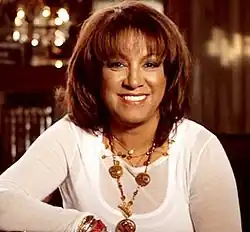 A woman in white clothing, standing in front of a red curtain and wearing gold jewelry.