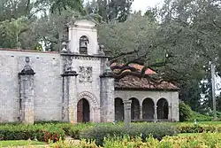 The corner of a stone church with hedges close to the viewer and tall trees in the background. We can see the gate of the church with a belfry on top and four arches of an extension covered by tiles.