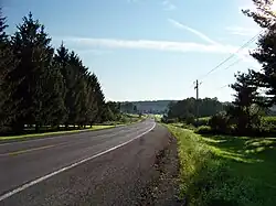 A two-lane highway heads through an area containing open fields and areas of trees. A large, tree-covered ridge is in the distant background.