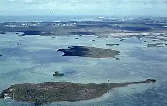 A large lake seen from above. The water is dark blue in some areas and light blue in others. Multiple islands are visible in the lake, as well as a shoreline in the distance with what appears to be vegetation and buildings.