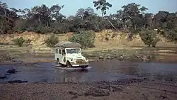 A photograph of a white vehicle with luggage on top driving across a river with green trees in the background all under a clear blue sky.