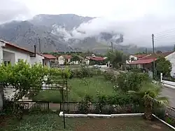 White house’s with red tile roofs surrounding a grassy area. A mountain is visible in the background with clouds/fog obscuring the rightmost portion of the mountain.
