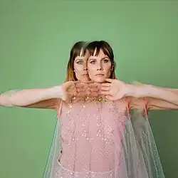 Oh Land is shown wearing a flowing, pink dress atop an identical clone of herself against a green background.