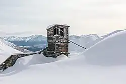 "A wooden building with slanted roofs and a cable coming out of one of the towers. Large, smooth drifts abut the building on one side, and only a three-story tower is fully exposed. In the background is a wide valley, with more mountains on the other side."