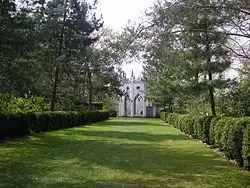 A colour photograph of grassy track lined with trees and hedges leading to a white gothic folly