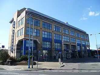 A four-storey modern yellow-brick building with many large blue windows on the facade. In the left hand corner, some of the building is cut out with a pillar supporting the structure and a glass structure inside. In the centre on the bottom floor is two entranceways, the left of which has a sign that reads "Potters Bar" above. There are also vertical signs that read "Potters Bar" in between the windows on each side of the entranceway.