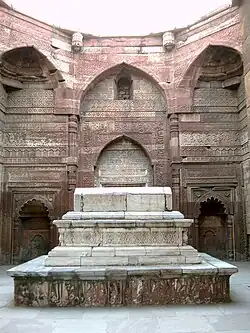 A color photograph of a white marble tomb placed on a square platform. Behind the tomb are tall red walls with Persian and Arabic calligraphy inscribed on them
