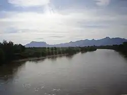 A wide flat river flows between tree-lined banks. Jagged hills rise in the distance.