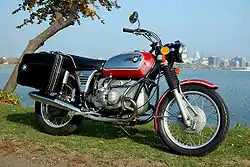 Red BMW R75/5 motorcycle fitted with panniers, parked on grass by a lake with a city in the background