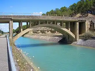 A bridge spans over a turquoise canal surrounded by dry terrain and trees.