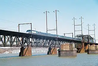 A high-speed passenger train crossing a multi-span truss bridge over a wide river. Masonry piers are visible near the bridge.