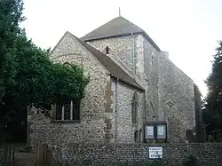 A flint rubble church with large stone quoins, behind a low flint wall. A three-light lancet window in the nearest wall is partly obscured by a tree in full leaf. A squat tower with a squared-off pyramidal roof stands in the middle. To its right is a blank-walled protrusion with a blocked entrance.