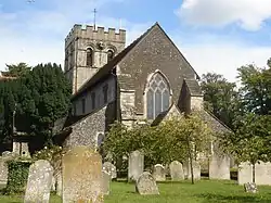 Rear view of a stone church with a castellated tower at the far end. The nearest side has very dark stone, two heavy buttresses and a three-light lancet window with trefoils. Trees surround the church on all sides, and there are several gravestones in front.