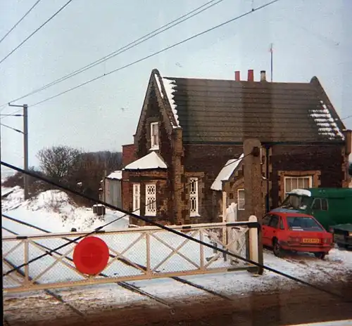 A small brick building with ornate windows situated to the right of two railway tracks. There is a level crossing barrier across the tracks, cars in front of the house, and overhead gantries above the track. There is no other visible evidence of a former station. The ground is snowy and the photo has a sepia tone.