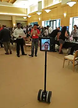 The image depicts the type of mobile telepresence device that would be used. It's on wheels, about 5 feet tall or shorter, and has a tablet device attached on top showing a mans face.