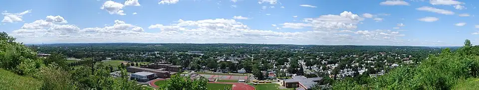 A wide panoramic shot begins and ends in bushes but is graced by two- and three-story city houses throughout most of the image. Front and center are three large schools featuring a track, tennis courts, and a football field. Behind the houses is a river flowing right to left.