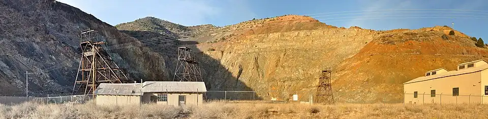 Panorama of fenced-off pit surrounded on three sides by rock walls. Three headframes and two buildings are within the enclosed area near the pit.