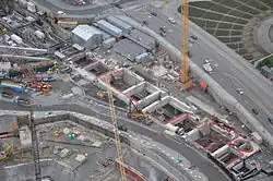 A construction site with several cranes and walls surrounding a long, rectangular hole in the ground.