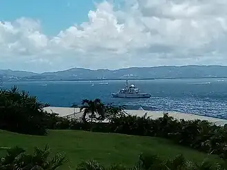 A body of water with a boat in it and tree-covered hills in the distance. In the forefront is the top of a grassy piece of land with bushes and trees, and some roofs or structures just beyond it.