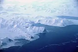 A photo taken from an aircraft of an artic bay, with dark and calm marine waters on the bottom of the image and glacial mountains covered in snow for the top half of the image.