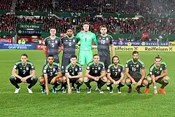 Photo of the Welsh national team posing before a game. There are four players standing and seven plays squatting, unevenly.