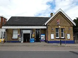 A brick building with an open entranceway below a canopy. On the right-hand side of the building there is a double arrow sign with "Winchmore Hill" written below. To the right of the entranceway there are ticket machines, newspaper stands, and a defibrillator.