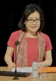 blurry image of Yuen Yuen Ang wearing a pink short-sleeved top and patterned scarf draped over her shoulders, in front of a desk with various items, including a computer keyboard and microphone, looking left of camera and appearing to speak.