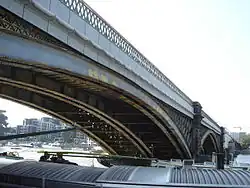 View from a barge of long arch above and barge passing beside, side of arches also visible with diamond-shaped painted balustrades. Backdrop of mid-storey buildings behind a tree-lined promenade.