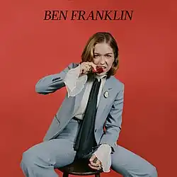 Cover image of "Ben Franklin": a woman wearing a blue suit is biting a rose while sat on a stool. The name of the song in all caps, "BEN FRANKLIN", is printed above her.