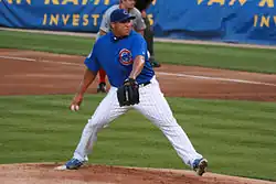 Full shot of pitcher in mid pitch. Pitcher is wearing a blue hat, a blue shirt with a Cubs logo and white pants