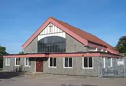 A low, grey stone-faced building behind a car park, with a security fence to the right. The front section (with a double entrance door) is wide and flat-roofed; behind this a red-tiled pointd roof rises to cover the main body of the church. All soffits are a weathered red colour. An 11-light window with some white panelling and a crucifix above dominates the façade.