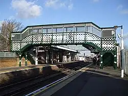 Two people walking on a green bridge with a white lattice running over a railway track and platform under a blue sky with white clouds