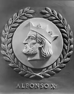 A dark-grey marble side-bust of Alfonso X. He has long hair, a bear, and a crown that tops his head. He is looking left, and leaves wrap around the circular engraving that the side-bust sits in. Below the work is the text "ALFONSO X".