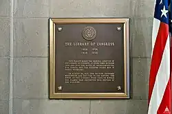 A black-ang-gold bronze plaque that reads: "THE FIRST LIBRARY OF CONGRESS 1802-1804 1810-1814 THIS TABLET MARKS THE ORIGINAL LOCATION OF THE LIBRARY OF CONGRESS. AT OTHER TIMES BETWEEN 1800 AND 1810 THE HOUSE OF REPRESENTATIVES, THE SENATE AND THE SUPREME COURT MET IN THESE QUARTERS. ON AUGUST 24, 1814 THE BRITISH CAPTURED WASHINGTON AND SET FIRE TO THE CAPITOL. THE BOOKS IN THE LIBRARY WERE USED TO KINDLE THE FLAMES THAT DESTROYED THIS SECTION OF THE BUILDING". To the right of the plaque is an American flag.