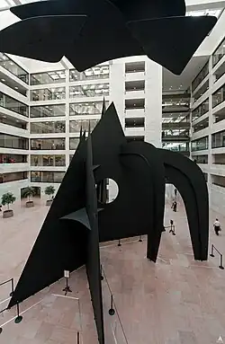A large black steel sculpture rises up from the ground. Behind the sculpture are rows of windows and catwalks, and on the floor are ropes to stop people from touching the statue. The second half of the statue hangs from the ceiling.