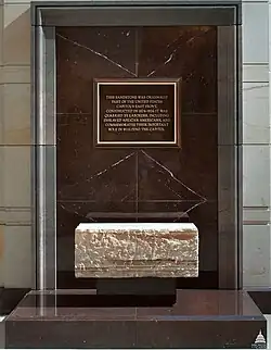 An enclosed glass box with a sandstone block on display. Behind the block is a black-and-gold plaque, with unreadable text.