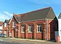 A dark red brick hall on a corner site, with adjoining houses in an identical style. The hall, partly obscured by a traffic light, has a pointed-arched entrance with a wooden door below a rectangular window frame with three lancets. To the right of the door, there are three plain windows between brick buttresses. Above the entrance is a steep roof perpendicular to the main roof.