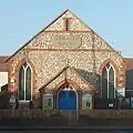 Front view of a tiny, plain chapel of irregular flintwork and red brick dressings. A dark stone plaque below a slit window near the roofline reads "WESLEYAN CHAPEL". An entrance porch is flanked by two wide, low-set windows with pointed arches.
