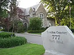 A close up picture of an address rock reading Hamilton Cemetery, 777 York Blvd.. The property has various bushes and trees and a large brick house in view.