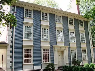 A three story colonial house with blue wood siding in front and brick walls on the sides.