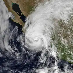 A photograph of a hurricane near the Pacific coast of Mexico; its eye is mostly obscured by clouds, and there is a large shield of clouds extending to the north and distant northeast of the center