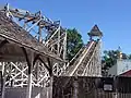 A historic roller coaster with white supports and a brown track with a steeple over the top of the left hill